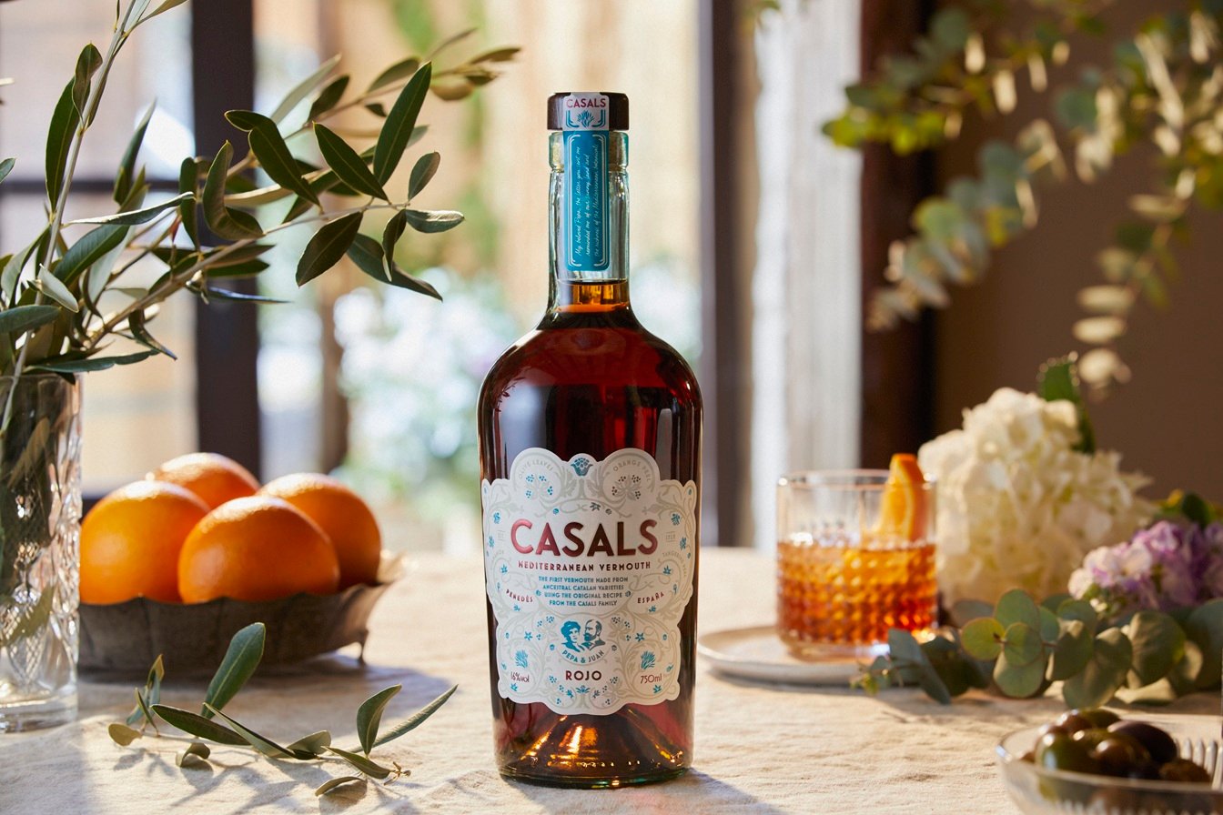 Botella de Casals Vermut Rojo sobre una mesa de aperitivo, con naranjas, aceitunas y una copa de vermut”.